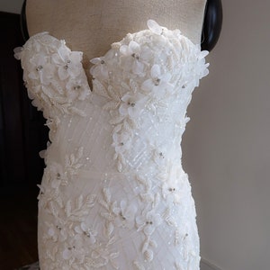 May include: A white strapless wedding dress with a fitted bodice and a flowing skirt. The bodice is adorned with delicate white flowers and sparkling beads.