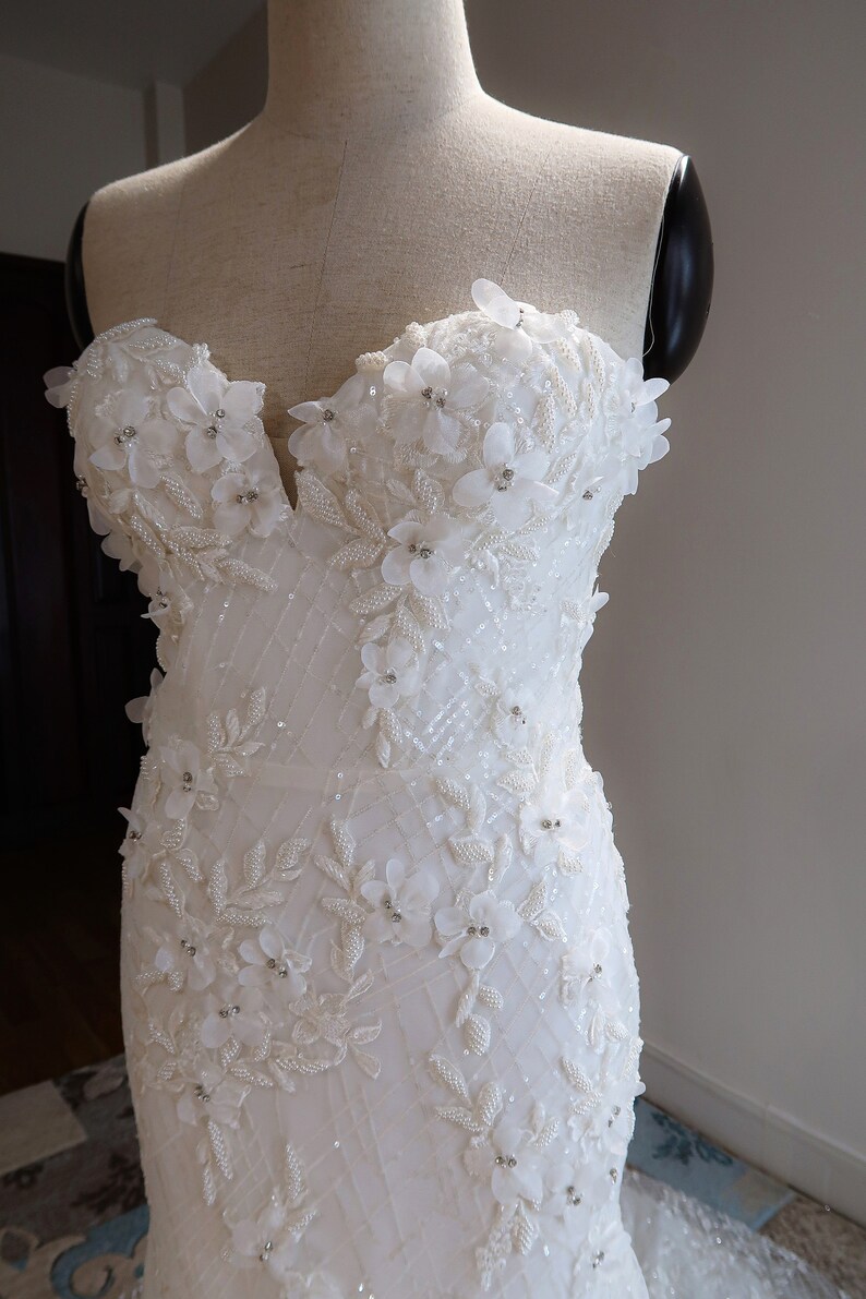 May include: A white strapless wedding dress with a fitted bodice and a flowing skirt. The bodice is adorned with delicate white flowers and sparkling beads.