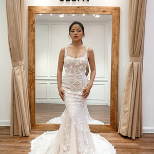 May include: A woman wearing a white wedding dress with floral lace detailing. The dress has a fitted bodice and a flowing skirt. The dress is being displayed in a store with a sign that reads "SELFA".