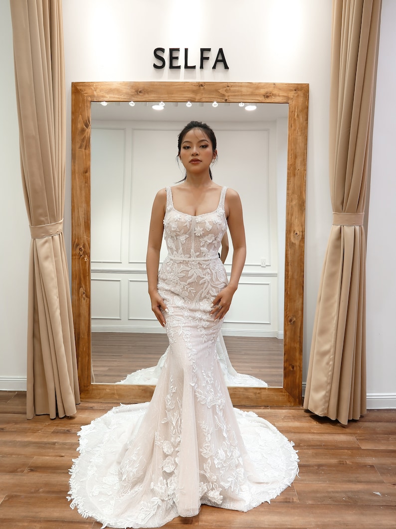 May include: A woman wearing a white wedding dress with floral lace detailing. The dress has a fitted bodice and a flowing skirt. The dress is being displayed in a store with a sign that reads "SELFA".