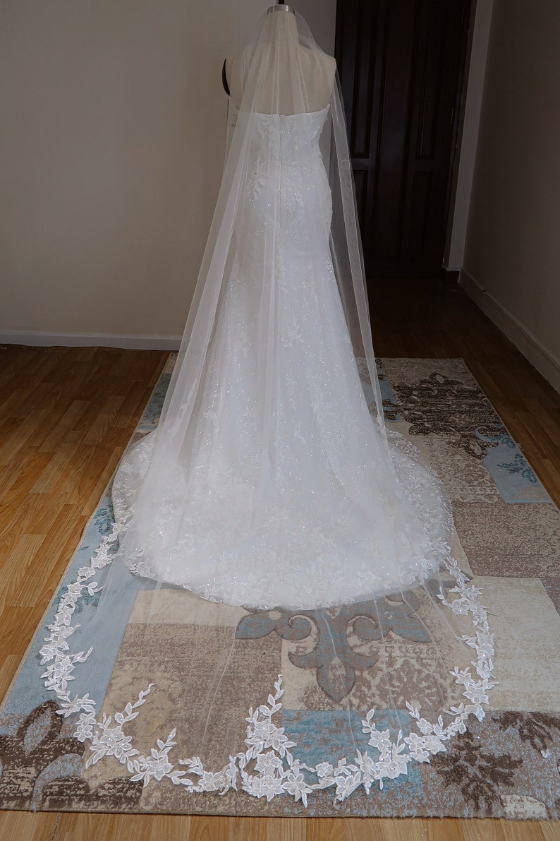 May include: A white bridal veil with a delicate floral lace trim. The veil is made of soft tulle and has a long, flowing design.