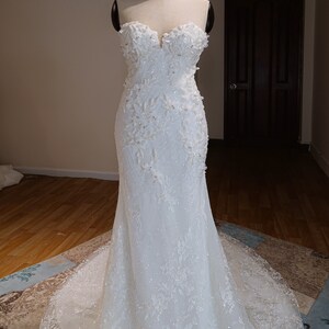 May include: A white strapless wedding dress with a fitted bodice and a flowing skirt. The dress is adorned with delicate floral lace and sequins.