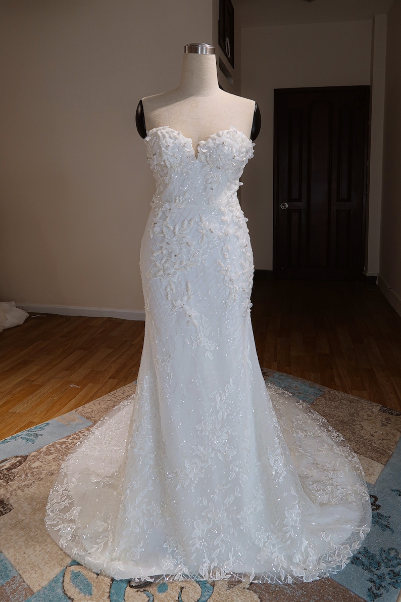 May include: A white strapless wedding dress with a fitted bodice and a flowing skirt. The dress is adorned with delicate floral lace and sequins.