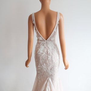 May include: A white and blush-colored wedding dress with a deep V-back and intricate floral lace detailing. The dress has a fitted bodice and a flowing skirt.