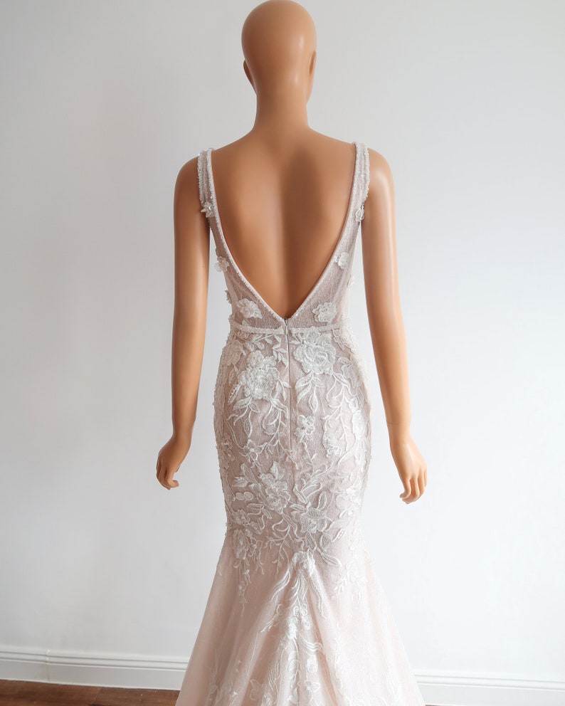 May include: A white and blush-colored wedding dress with a deep V-back and intricate floral lace detailing. The dress has a fitted bodice and a flowing skirt.