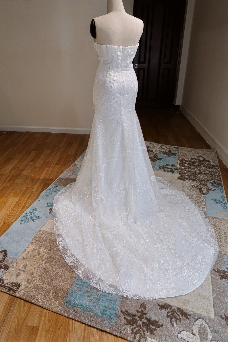 May include: A white strapless wedding dress with a fitted bodice and a flowing skirt. The dress is adorned with delicate lace and sequins.