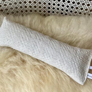 Könnte beinhalten: Ein weißes, rechteckiges Objekt aus gewebtem Stoff mit elastischen Bändern an jedem Ende, das auf einer cremefarbenen, flauschigen Oberfläche liegt. Das Objekt hat ein strukturiertes Muster und ein kleines Etikett mit Text.