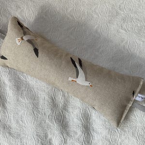 Può includere: Un fermaporta beige con una fodera in tessuto bianco con un motivo di gabbiani in volo. Il fermaporta ha un'impugnatura ad anello e un piccolo cartellino con la parola "Gabbiano" stampata su di esso.