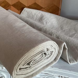 Può includere: Un grande rotolo di tessuto color beige chiaro naturale con una trama visibile. Il materiale è arrotolato strettamente su un lato, con una parte del tessuto srotolata e distesa in piano per rivelare la trama consistente e il tono neutro.