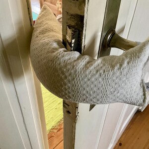 Könnte beinhalten: Ein hellgrauer Türluftstopper aus strukturiertem Stoff, der um einen Türgriff gewickelt ist. Der Stopper soll Zugluft blockieren und Geräusche reduzieren. Ein kleines weißes Etikett mit Text ist angebracht.