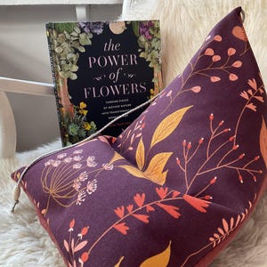 Peut inclure: Un pouf prune avec un motif floral avec des feuilles orange et jaunes et des baies rouges. Le livre "The Power of Flowers" est visible en arrière-plan, posé sur une chaise blanche.