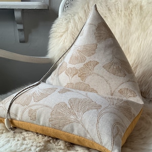 Può includere: Un cuscino decorativo triangolare con un tessuto di lino beige con un motivo ripetuto di foglie di ginkgo stilizzate di colore marrone chiaro. Il bordo inferiore è giallo senape e un dettaglio di corda è attaccato.