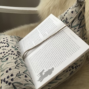 Può includere: Un libro appoggiato su un paio di cuscini di lino con un motivo floreale in tonalità di verde e viola. Il libro è aperto a una pagina con testo in tedesco. Uno spago viene utilizzato per tenere il libro aperto.