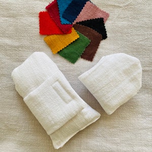 Könnte beinhalten: Zwei weiße Stoffbeutel mit Klappenverschluss. Ein Fächer mit Stoffproben in verschiedenen Farben, darunter Rot, Blau, Grün und Braun, wird auf einer weißen, strukturierten Oberfläche präsentiert.