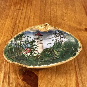 May include: Hand-painted seashell featuring a lighthouse scene. The artwork depicts a white lighthouse with a red roof, surrounded by green trees and a blue sky. The shell has a gold-colored rim and rests on a wooden surface.