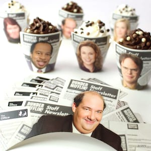 May include: A collection of cupcakes decorated with chocolate frosting and sprinkles. Each cupcake is wrapped in a paper liner featuring a character from the TV show "The Office" and the text "Dunder Mifflin Staff Newsletter".