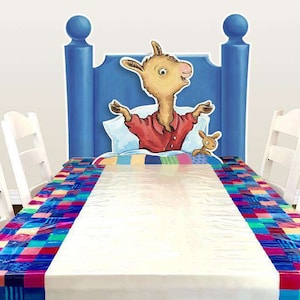 May include: A white tablecloth with a colorful checkered border. The tablecloth is laid on a white table with four white chairs. A cardboard cutout of a llama in a bed is behind the table.