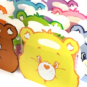 May include: A set of colorful cardboard party favor boxes featuring Care Bears characters. The boxes are shaped like bear faces with different colored backgrounds and have a handle on top.