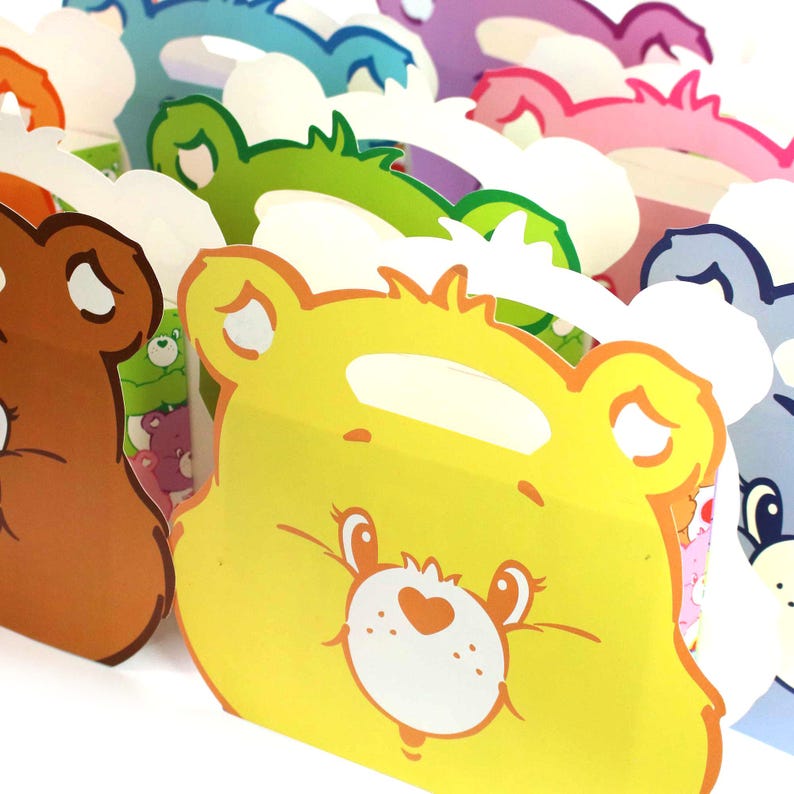 May include: A set of colorful cardboard party favor boxes featuring Care Bears characters. The boxes are shaped like bear faces with different colored backgrounds and have a handle on top.