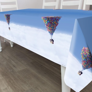 Op de afbeelding: Een rechthoekig tafelkleed met een herhalend patroon van een klein huisje dat door een grote tros veelkleurige ballonnen de lucht in wordt getild. Het ontwerp is geplaatst tegen een helderblauwe lucht met witte wolken aan de onderrand.