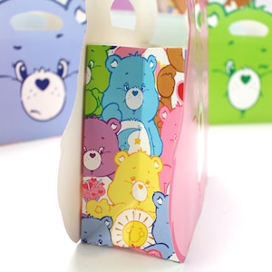May include: Pink and white paper gift bag with a handle. The bag is decorated with a pattern of colorful Care Bears.
