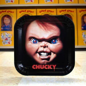 May include: Black square plate featuring a close-up of Chucky's face with red hair, blue eyes, and a sinister grin. The word "CHUCKY" is printed in red at the bottom. Background includes other Chucky-themed items.