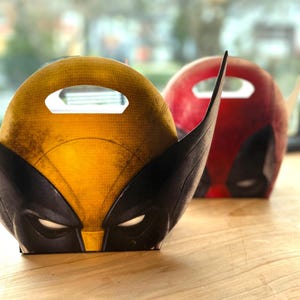 May include: Two character-themed gift boxes. One is yellow and black, resembling Wolverine's mask, and the other is red, similar to Deadpool's mask. Both have a handle and eye cutouts.