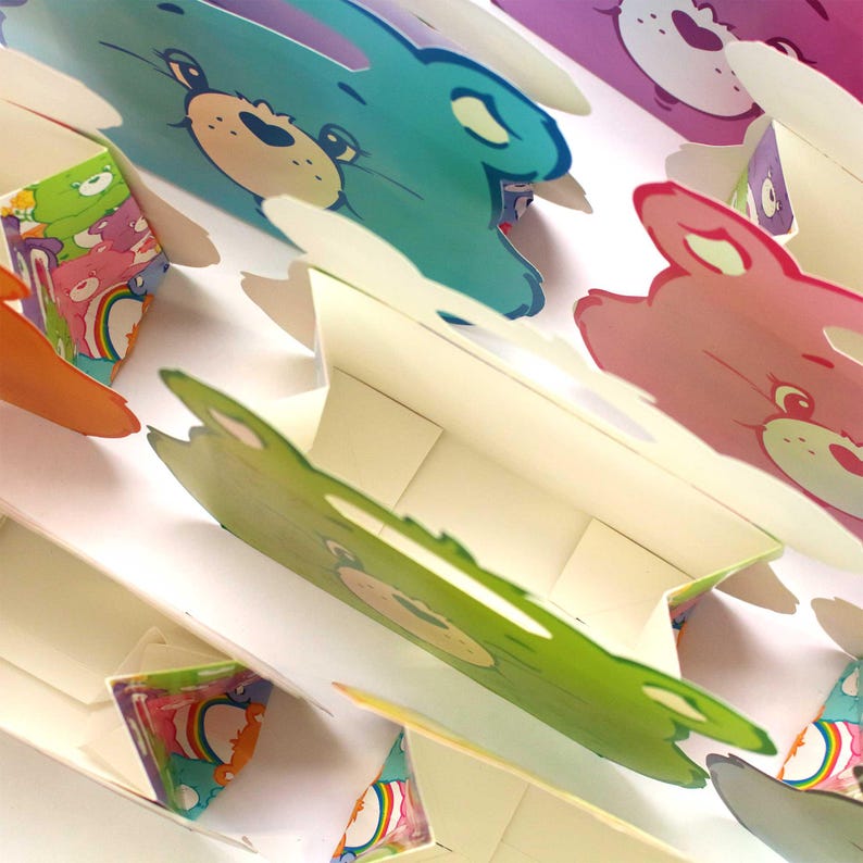 May include: A set of colorful Care Bear-themed gift bags. Each bag features a different Care Bear character with a rainbow design. The bags are made of sturdy cardboard and have a handle for easy carrying.