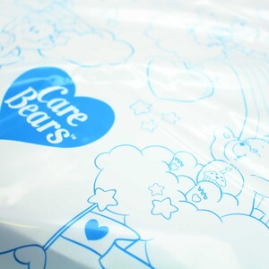 May include: A white plastic bag with a blue heart-shaped logo that says "Care Bears" in white text. The bag has a blue outline of a Care Bear holding a flag with a heart on it.