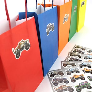 May include: A row of colorful gift bags in red, blue, orange, green, and yellow, each adorned with a monster truck sticker. Several sheets of monster truck stickers are in the foreground. The bags have white interiors and matching handles.