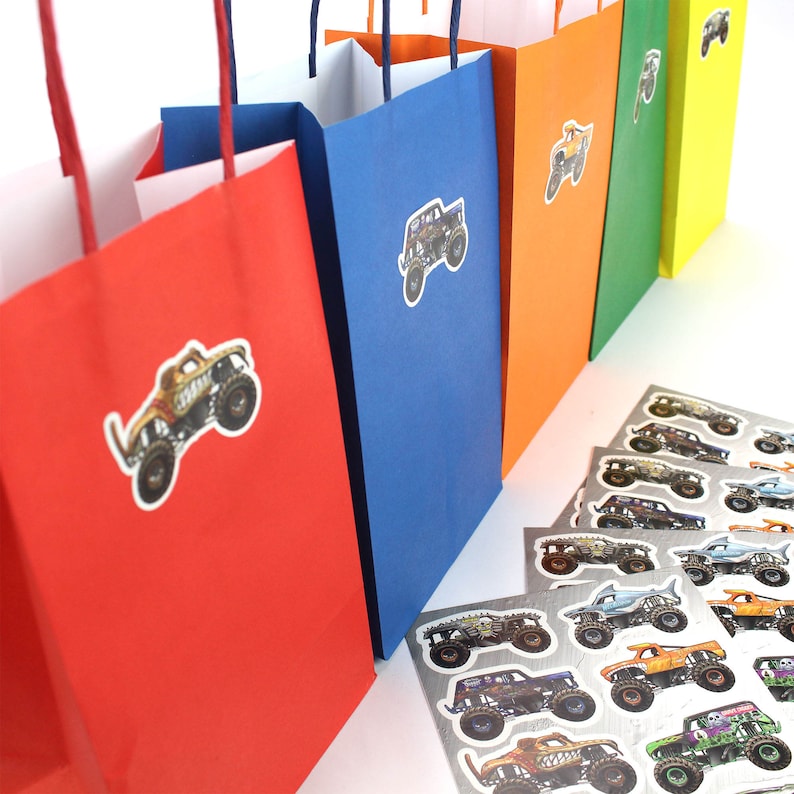 May include: A row of colorful gift bags in red, blue, orange, green, and yellow, each adorned with a monster truck sticker. Several sheets of monster truck stickers are in the foreground. The bags have white interiors and matching handles.