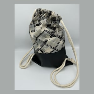 Könnte beinhalten: Ein Rucksack mit Kordelzug, mit schwarzem Boden und einem oberen Bereich mit einem Karomuster mit Sternakzenten in Schwarz, Grau und Weiß. Die Tasche hat dicke, cremefarbene Seilriemen.