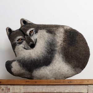 Op de afbeelding: Een grijs en wit vilten wolvenbeeld dat op een witgeschilderde houten tafel ligt. De wolf heeft een realistisch uiterlijk met zwarte ogen en een gekrulde staart.