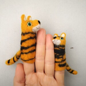 Op de afbeelding: Twee vingerpoppen in de vorm van tijgers. De grotere tijger is oranje en zwart met groene ogen. De kleinere tijger is oranje en zwart met groene ogen. Beide tijgers dragen een glimlach.