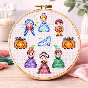 Op de afbeelding: Een borduurring met kruissteek met een sprookjesthema. Het ontwerp bevat afbeeldingen van een fee, een prinses, een prins, pompoenen, een glazen muiltje en andere personages. De borduursels zijn geplaatst in een houten ring.