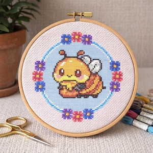 Könnte beinhalten: Ein Kreuzstich-Stickrahmen mit einem Cartoon-Bienen-Design. Die Biene ist gelb und orange, umgeben von einem blauen Kreis und einem floralen Rand aus rosa, lila und blauen Blumen. Der Rahmen ist aus Holz.