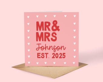 May include: A pink wedding card with the text "MR & MRS Johnson EST. 2025" in red. The card is decorated with small white hearts around the border and sits on a brown envelope. A perfect keepsake for a wedding.