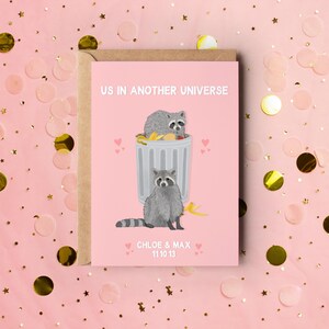 A pink greeting card with an illustration of two raccoons and the text ''Us In Another Universe"
