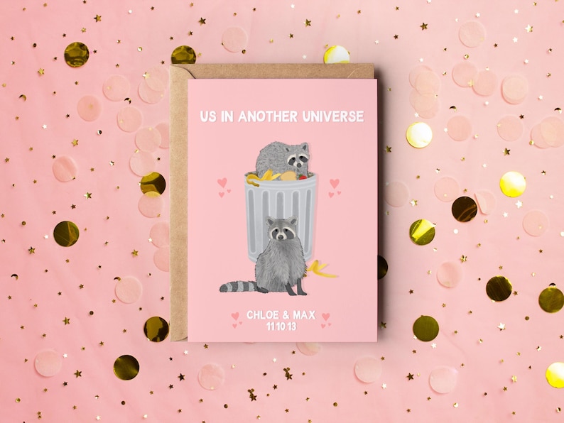 A pink greeting card with an illustration of two raccoons and the text ''Us In Another Universe"