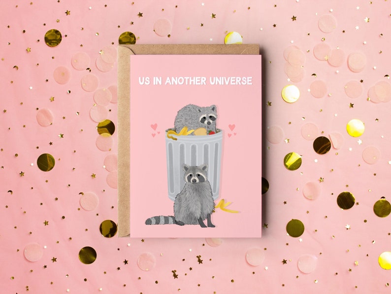 A pink greeting card with an illustration of two raccoons and the text ''Us In Another Universe"