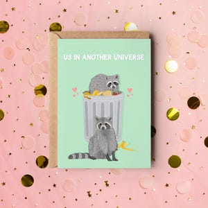 A green greeting card with an illustration of two raccoons and the text ''Us In Another Universe"