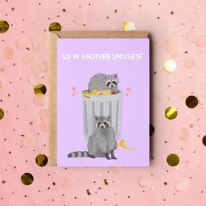 A purple greeting card with an illustration of two raccoons and the text ''Us In Another Universe"
