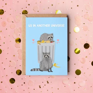 A blue greeting card with an illustration of two raccoons and the text ''Us In Another Universe"