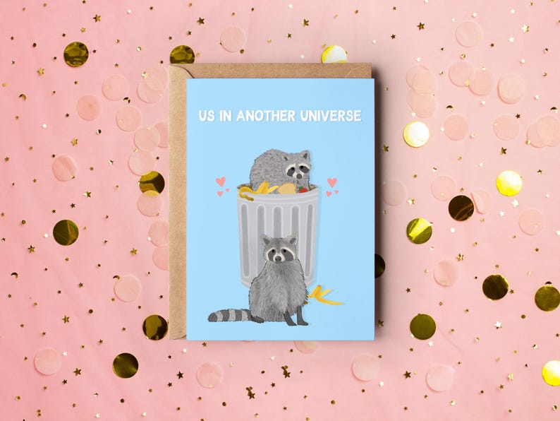 A blue greeting card with an illustration of two raccoons and the text ''Us In Another Universe"