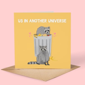 A yellow greeting card with an illustration of two raccoons and the text ''Us In Another Universe"