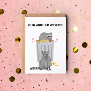 A white greeting card with an illustration of two raccoons and the text ''Us In Another Universe"
