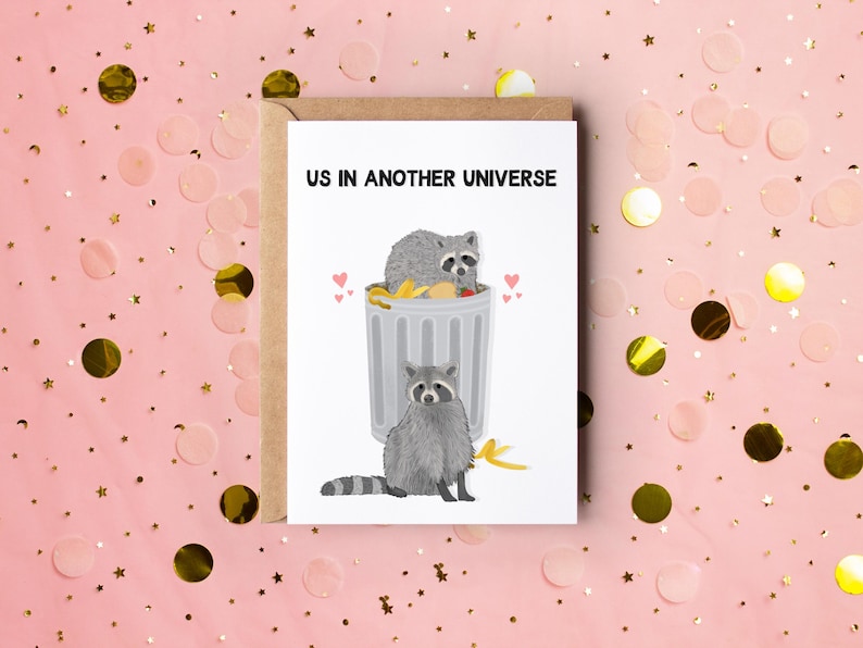 A white greeting card with an illustration of two raccoons and the text ''Us In Another Universe"