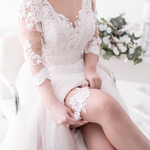 Könnte beinhalten: Eine Braut in einem weißen Spitzen-Hochzeitskleid mit V-Ausschnitt und transparenten Ärmeln, sitzt und justiert einen weißen Spitzen-Strumpfhalter an ihrem Bein. Der Hintergrund zeigt Blumendekorationen und Grün, was eine romantische Atmosphäre schafft.