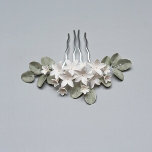 May include: A silver hair comb adorned with white flower blossoms and sage green leaves. The floral arrangement includes small, delicate flowers and larger blossoms, creating a natural, elegant design. This hair accessory is ideal for weddings or special occasions.