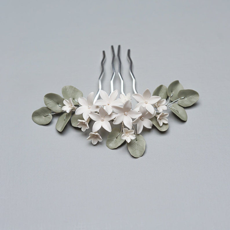 May include: A silver hair comb adorned with white flower blossoms and sage green leaves. The floral arrangement includes small, delicate flowers and larger blossoms, creating a natural, elegant design. This hair accessory is ideal for weddings or special occasions.